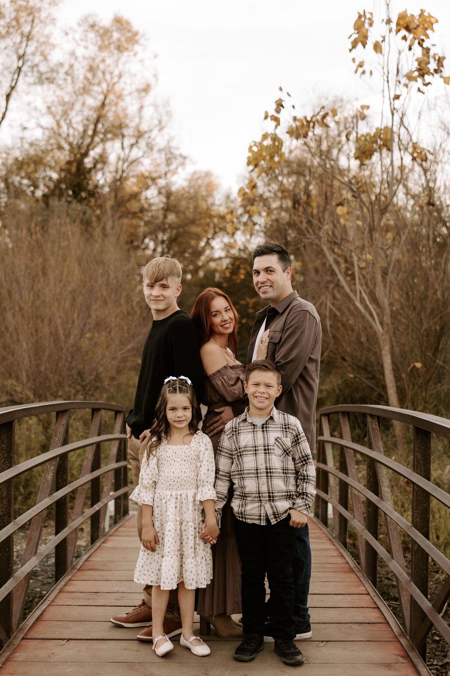 A family poses together on a wooden bridge in an autumn setting with trees in the background.