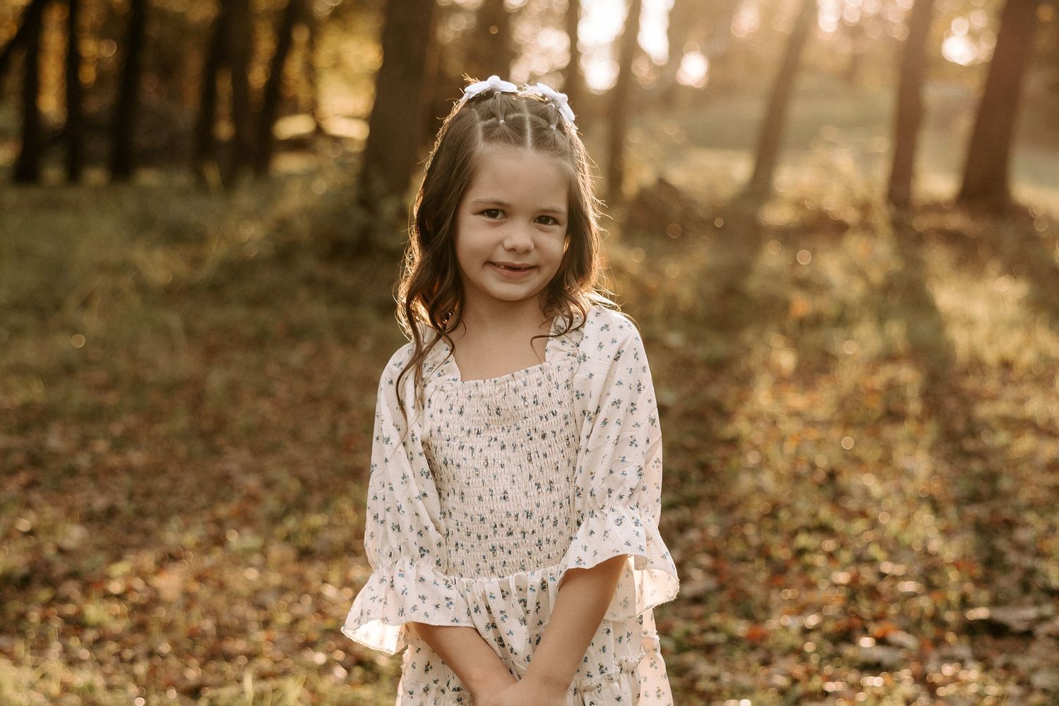 A young person in a white lace dress poses in a sunlit autumn forest with fallen leaves on the ground.