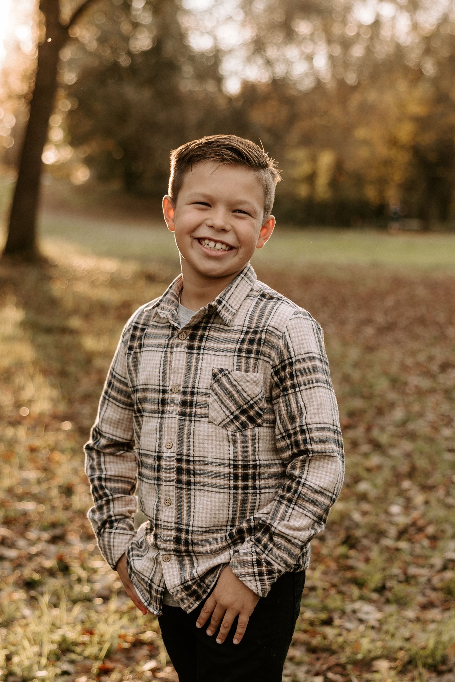 A young child in a plaid flannel shirt poses outdoors among fallen autumn leaves during golden hour.