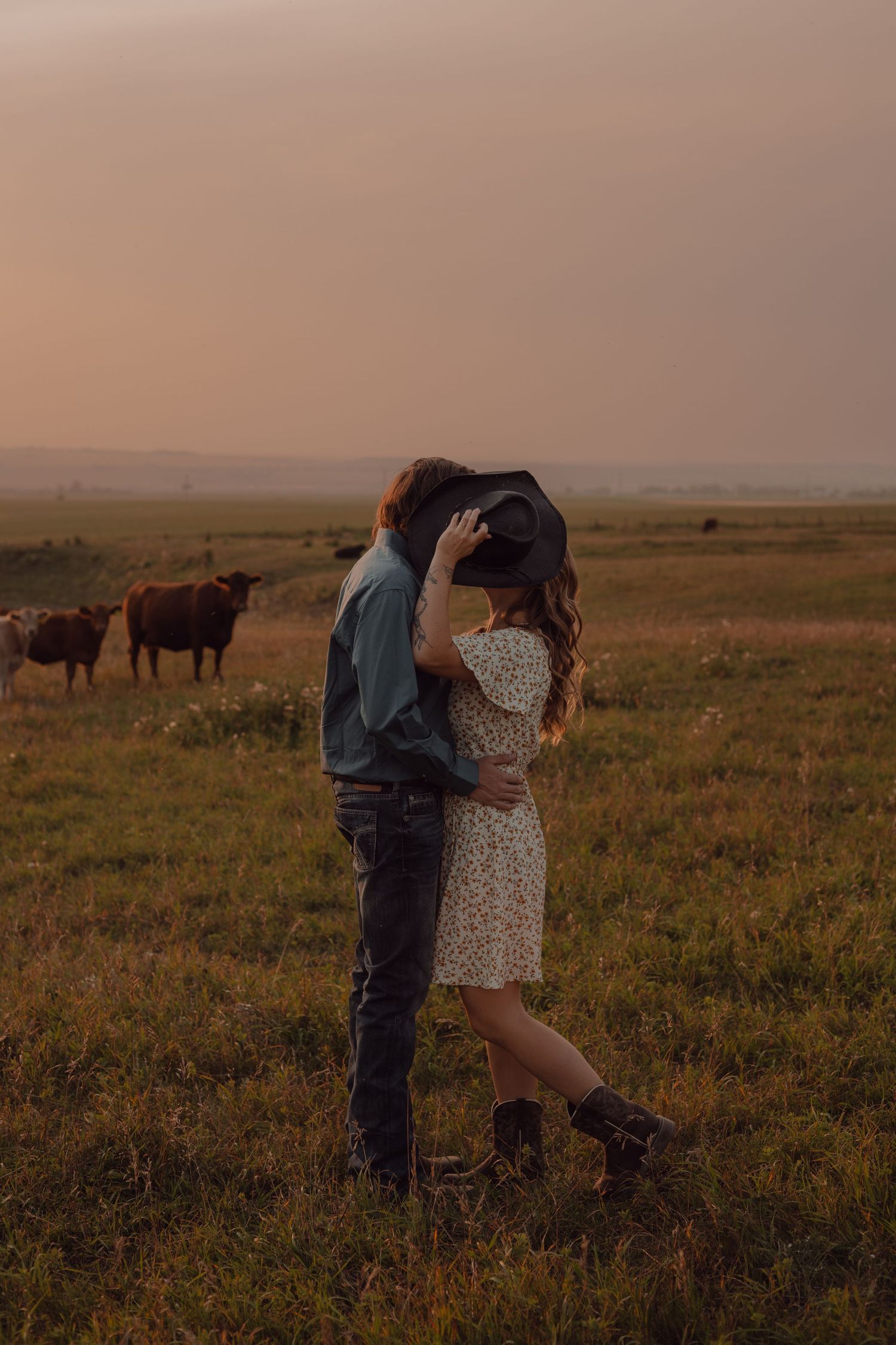 A warm couple's session in the farmland heart of Airdrie, Alberta. Overlooking the pasture, the couple sneak a kiss behind her cowboy hat.