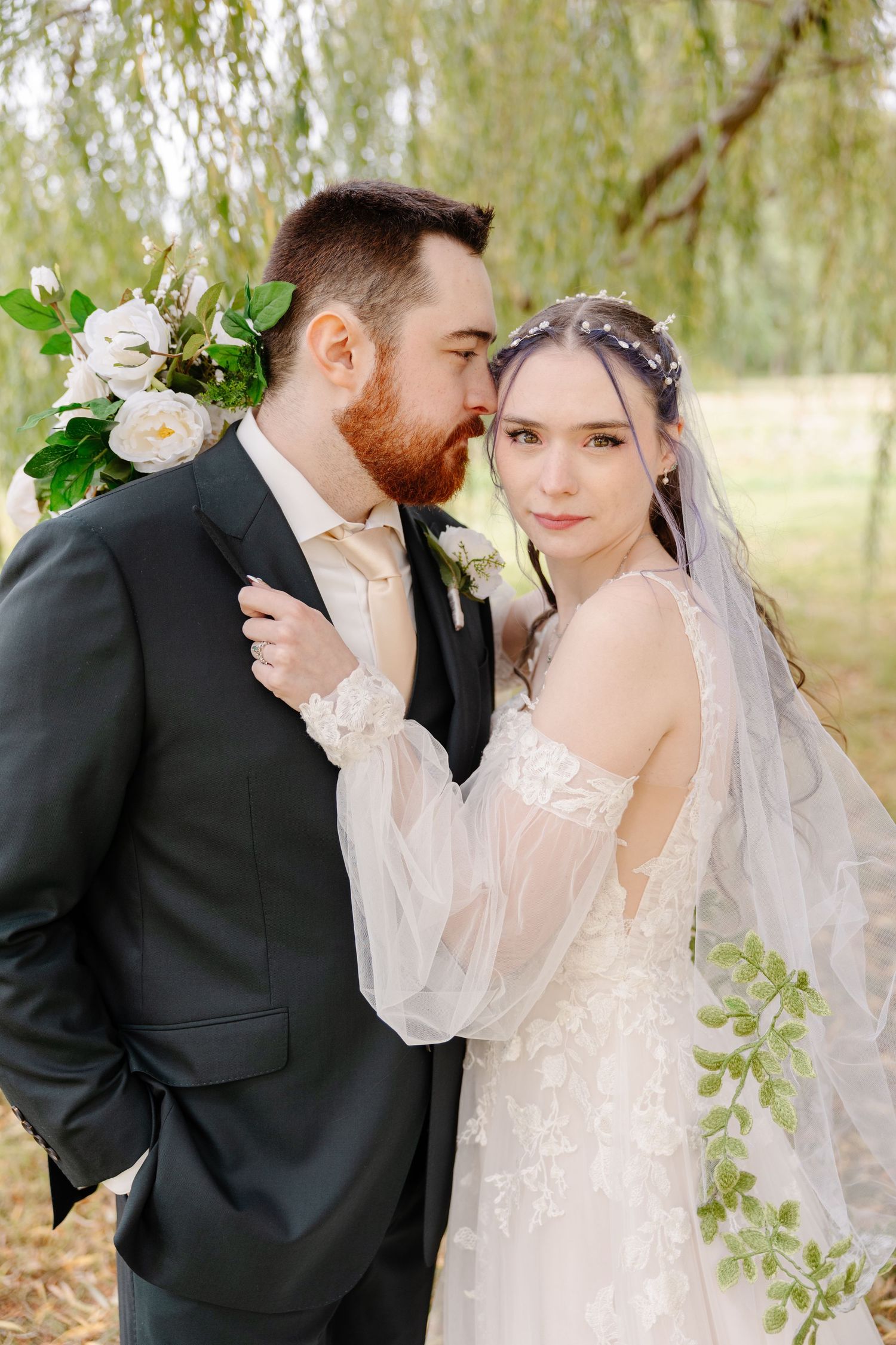 A romantic wedding couple shares intimate moments under willow trees in a dreamy outdoor setting.