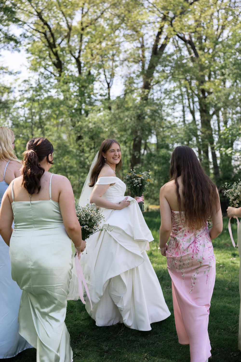 A group of women in pink and white dresses walk together through a wooded area on a sunny day.