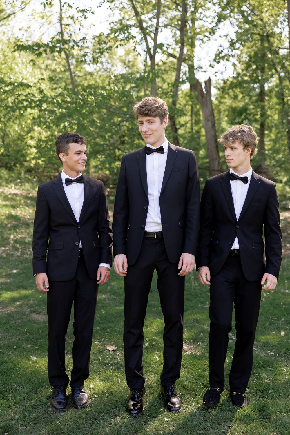 Three groomsmen in black tuxedos and white shirts stand together on grass in a natural outdoor setting.