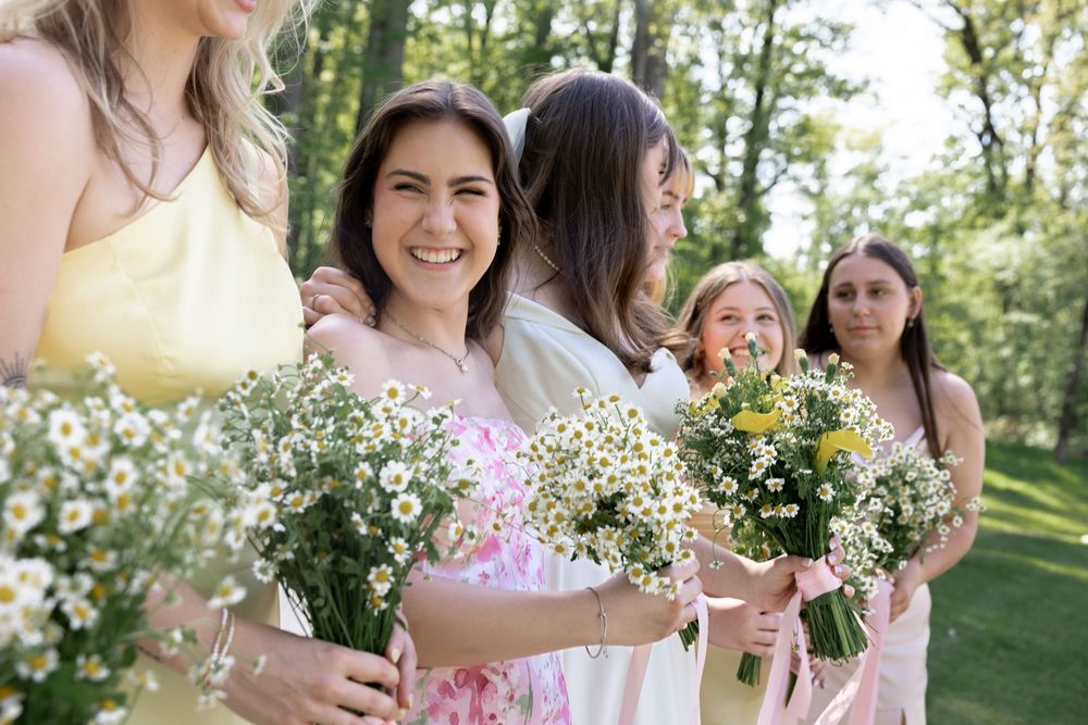 A wedding party stands outdoors holding delicate bouquets of pink and white flowers in a sunlit garden setting.