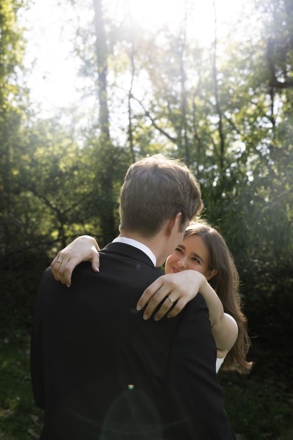 A couple shares an intimate embrace outdoors with sunlight streaming through trees in the background.