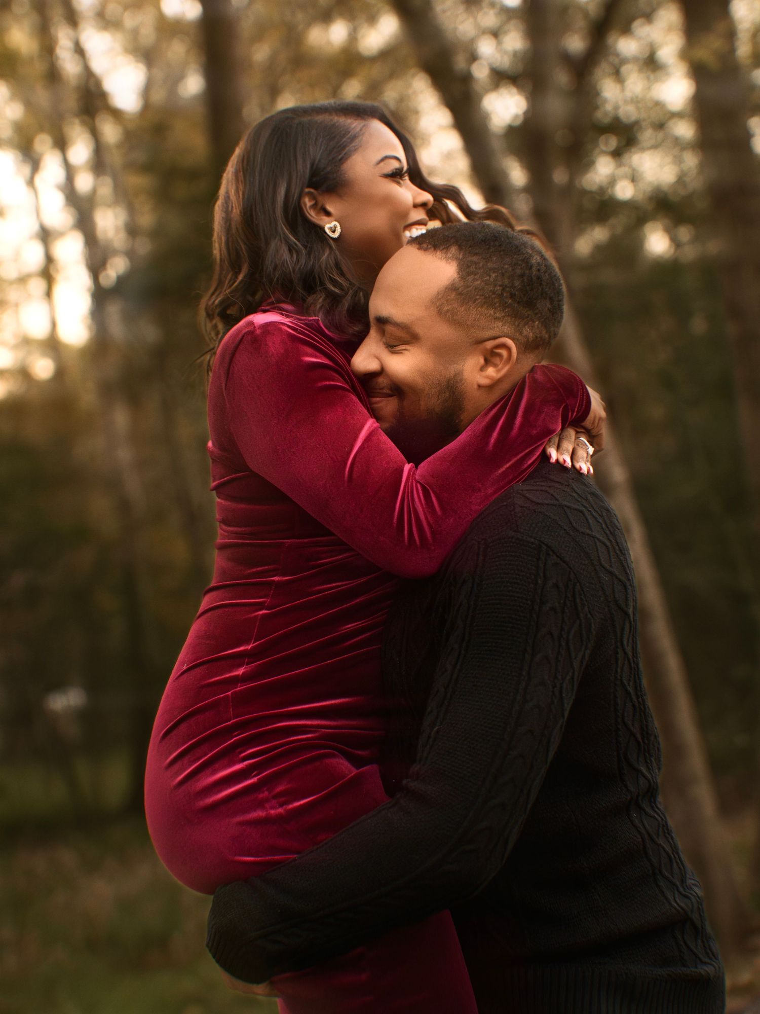 A romantic couple embraces outdoors at sunset, with the woman wearing a burgundy velvet dress and the man in black.