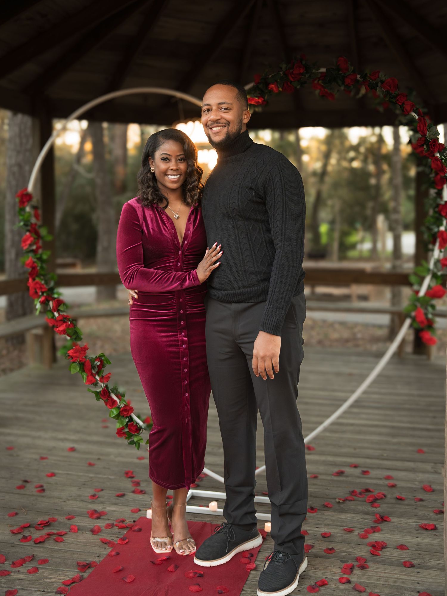 A couple poses under a floral arch with red roses, the woman in burgundy velvet and man in black casual wear.
