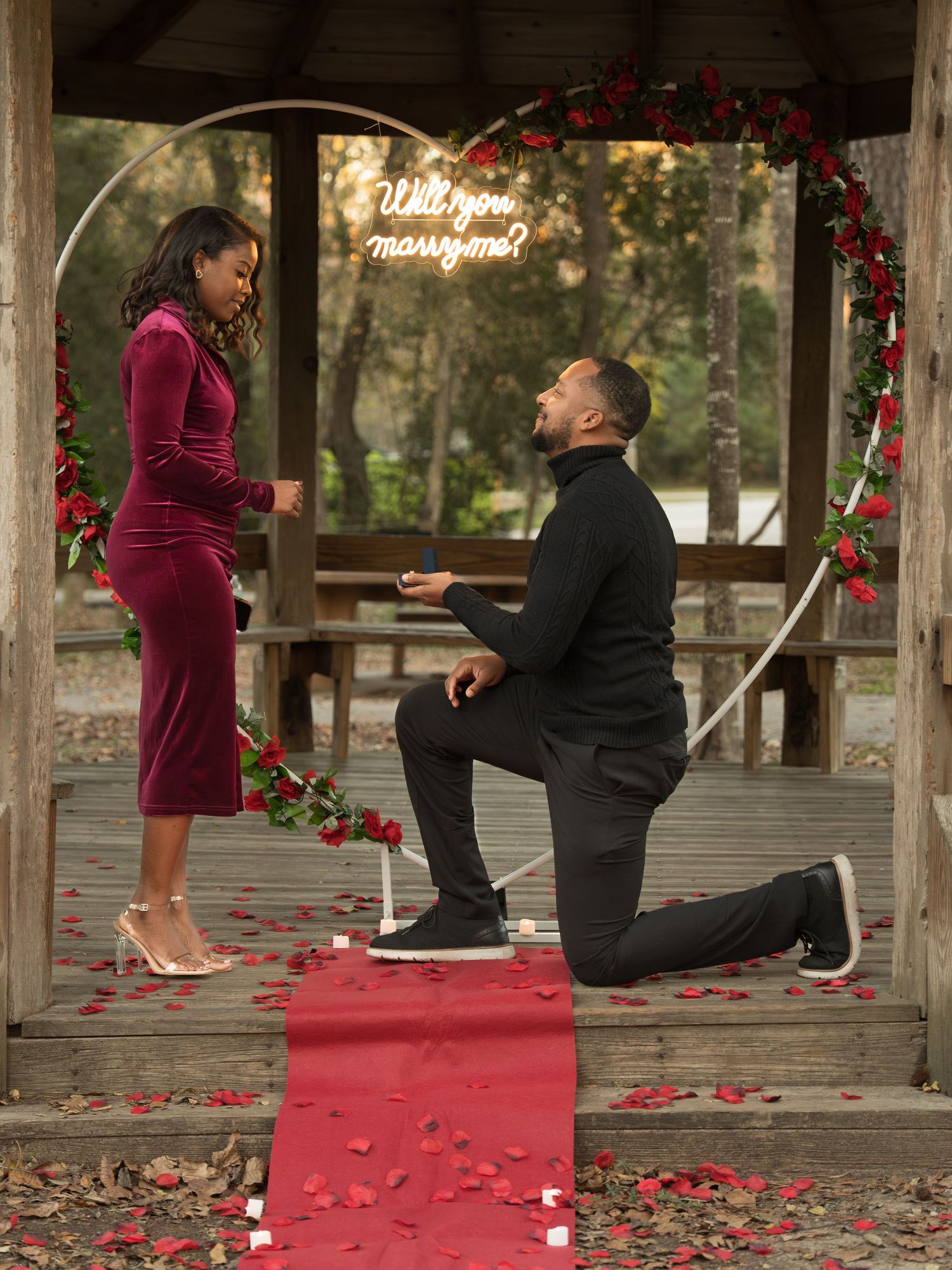 A romantic marriage proposal scene on a wooden deck decorated with scattered red rose petals.