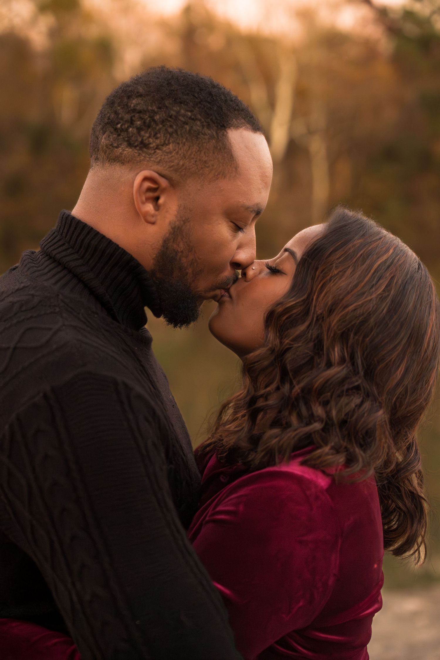 Romantic couple sharing intimate moments at sunset in a park wearing burgundy and black outfits.