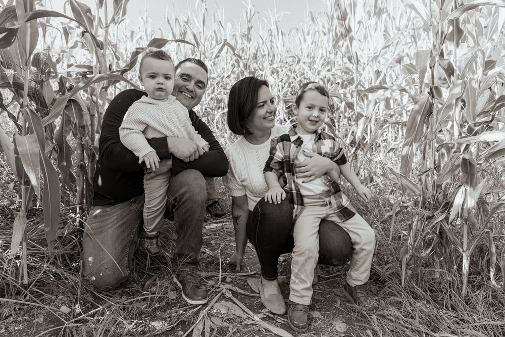 Black and white photo of a family of four posing together in a rustic cornfield setting.