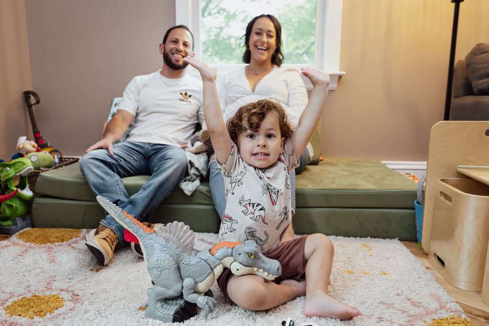A cheerful family relaxes indoors as their curly-haired toddler plays with a dinosaur toy.