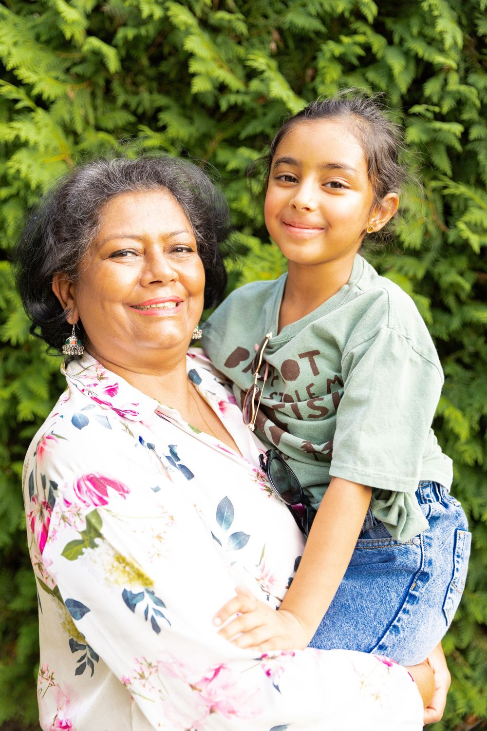Smiling grandmother holding young granddaughter outdoors in front of lush green hedges.