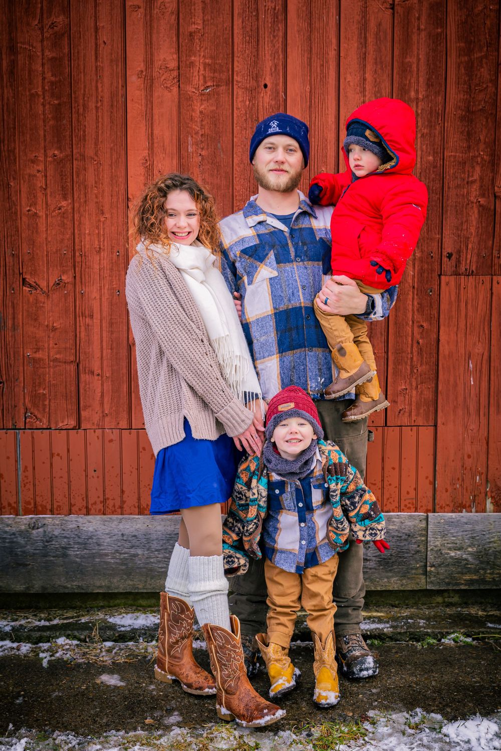 Young family of four posing in winter outfits against a rustic red barn wall in snowy weather.