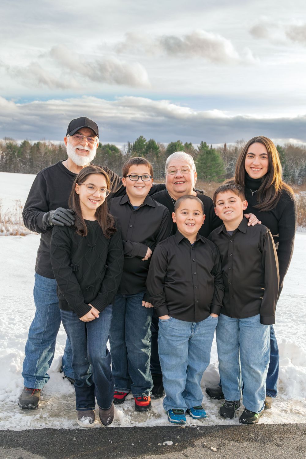 Extended family of seven in matching black shirts posing outdoors in snowy winter landscape.
