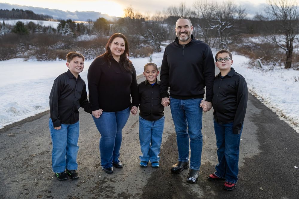 Family of five in black outfits holding hands on a snowy path at sunset in winter.