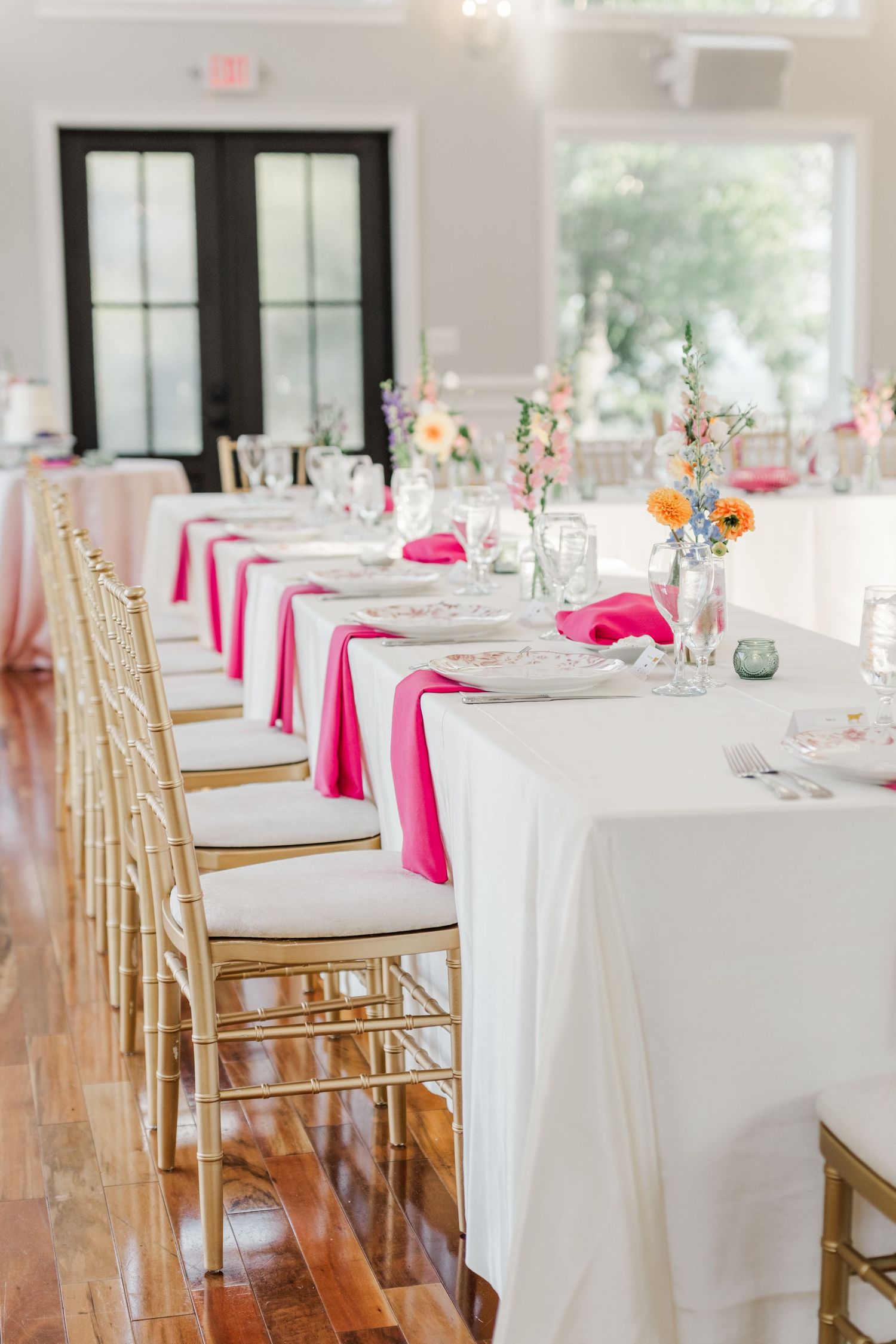 Elegant wedding reception table setting with white linens, pink napkins, and gold chiavari chairs in a bright venue.