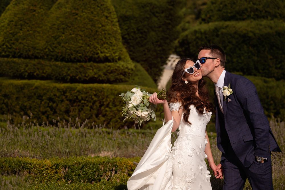 A couple shares a romantic moment in their wedding attire against a backdrop of manicured garden hedges.