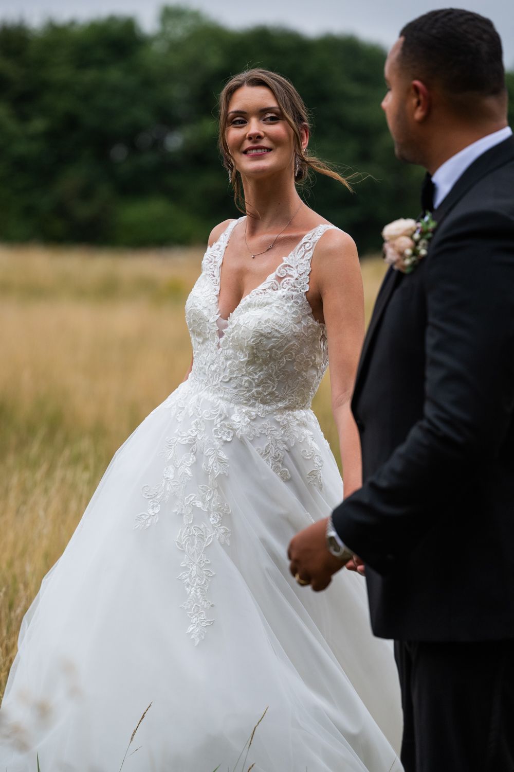 A bride in a white lace ballgown stands with her groom in a field during their outdoor wedding ceremony.