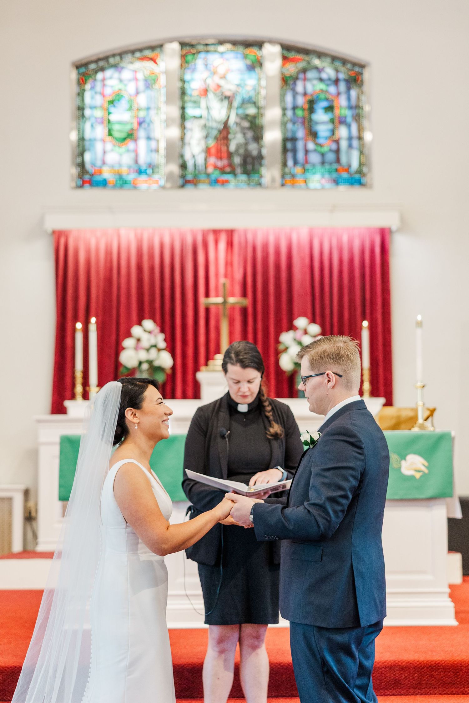 A wedding ceremony takes place in a church with stained glass windows and red carpet, with the couple exchanging vows.