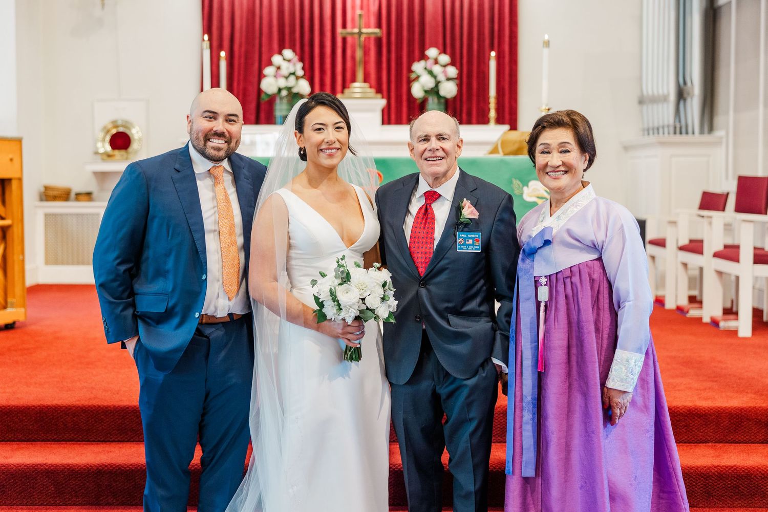 Wedding party poses together in formal attire with red carpet and church backdrop.
