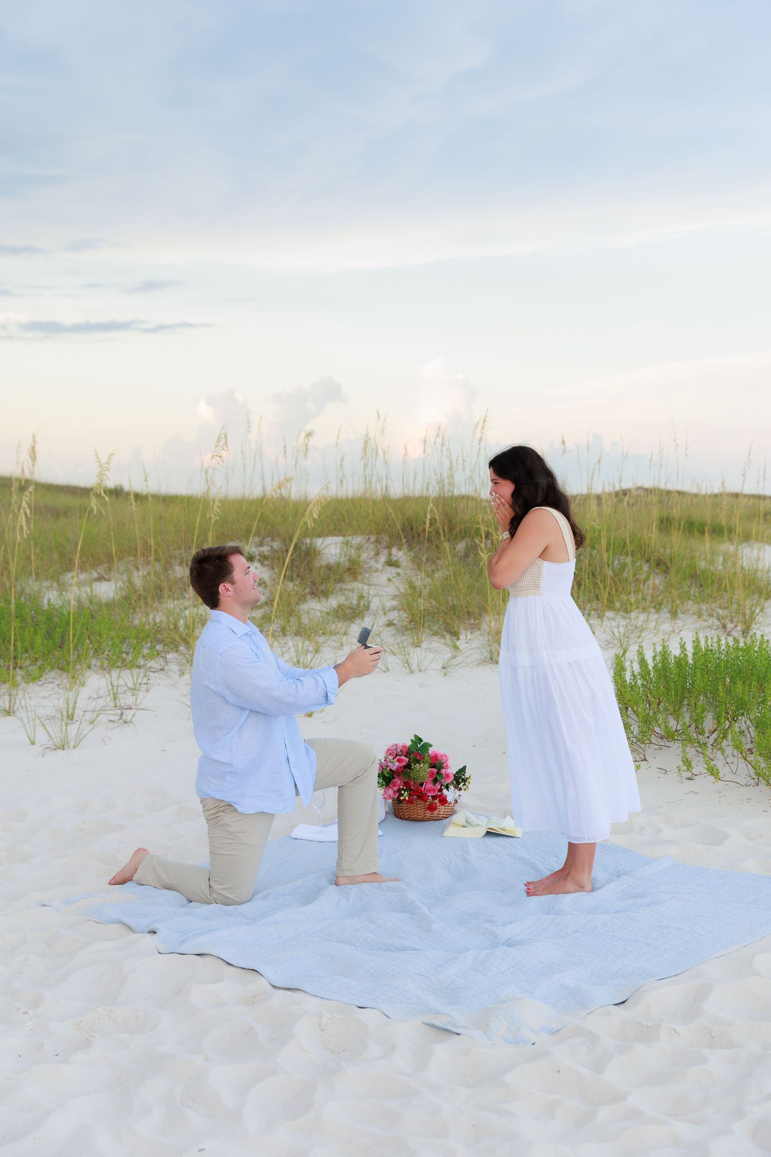 A surprise proposal moment captured on a beach blanket with flowers and coastal scenery at sunset.