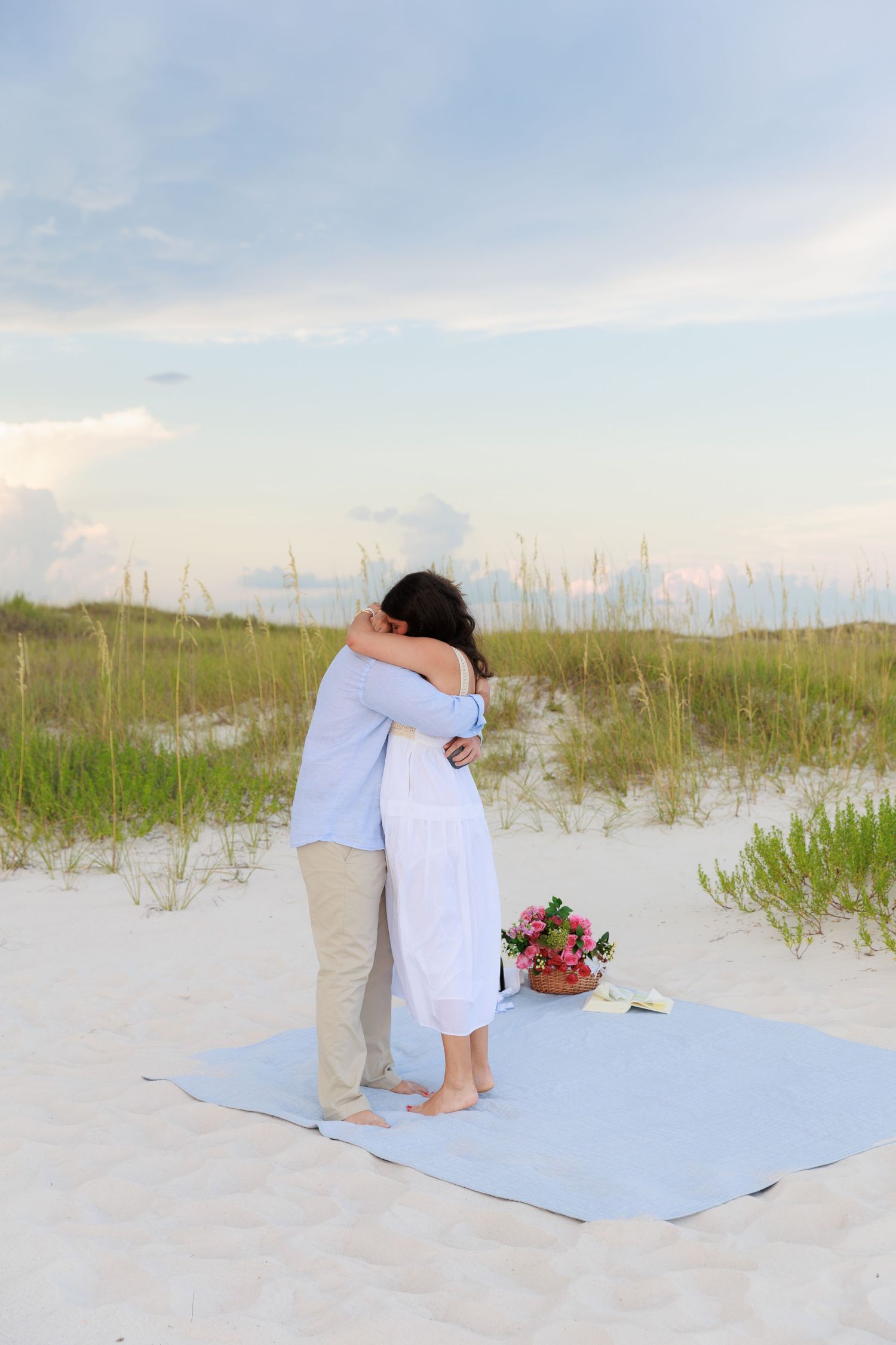 A romantic beach proposal scene unfolds on a light blue blanket with sand dunes and sea grass in the background.