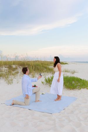 A romantic beach proposal scene with a blanket laid out on white sand dunes at sunset.