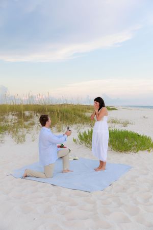 A couple shares an intimate moment during a beach proposal with ocean waves and dunes in the distance.