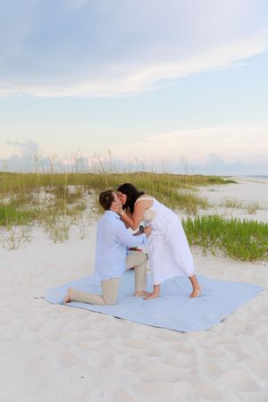 A romantic beach proposal scene unfolds on a white sandy beach with sea oats and gentle waves in the background.