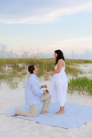 A couple shares an intimate moment during a beach proposal on a light blue blanket surrounded by coastal dunes.