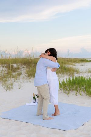 Two people embrace on a blue blanket after a beachside proposal with coastal vegetation in the background.