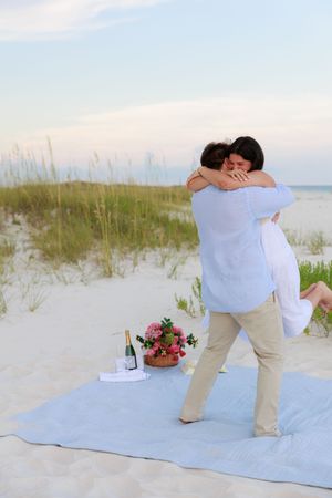 A joyful celebration embrace on a beach blanket with champagne and flowers nearby.