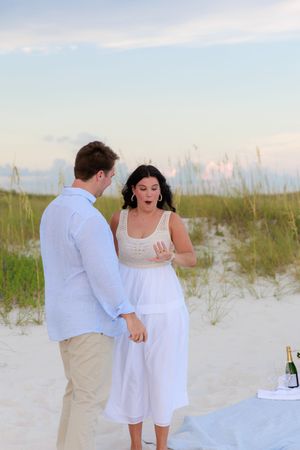 A couple dances playfully together on a white sandy beach with sea oats and dunes in the background at sunset.