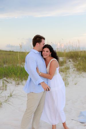 A romantic couple in white and blue outfits sharing an intimate moment on a beach with sea grass.