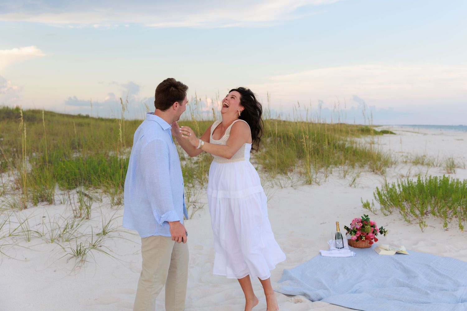Romantic couple in white outfits walking and dancing on a sandy beach at sunset with grass in the foreground.