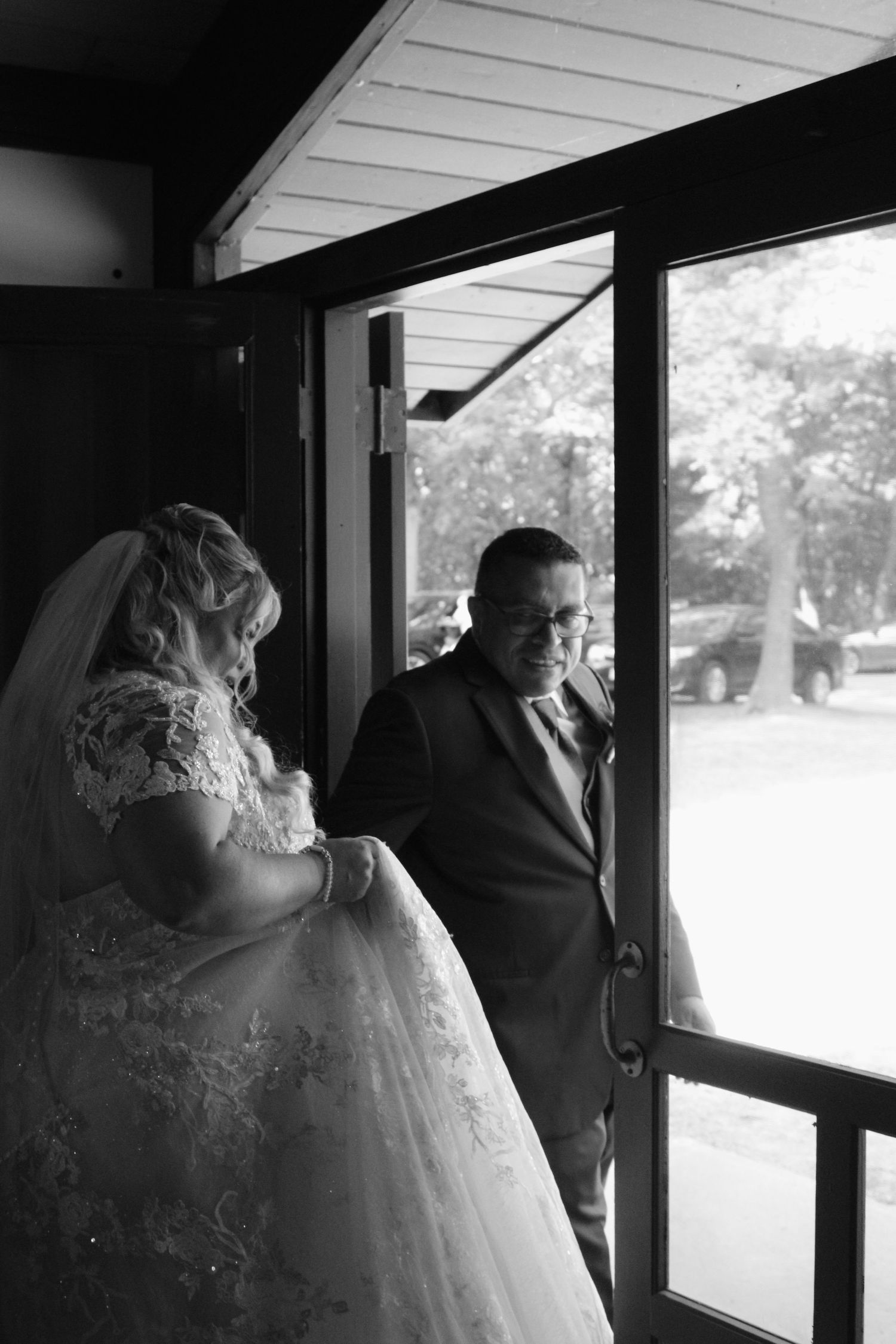 A black and white photo of a couple in wedding attire standing by a window in contemplative moment.