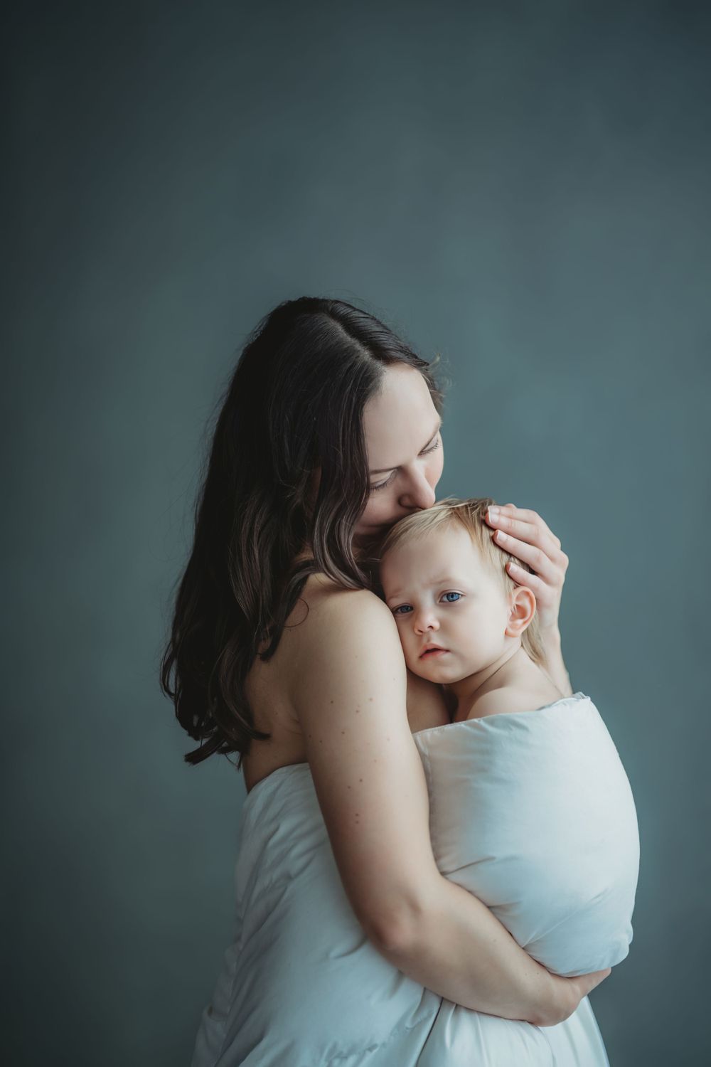 A tender moment between parent and baby wrapped in white fabric against a moody teal background.