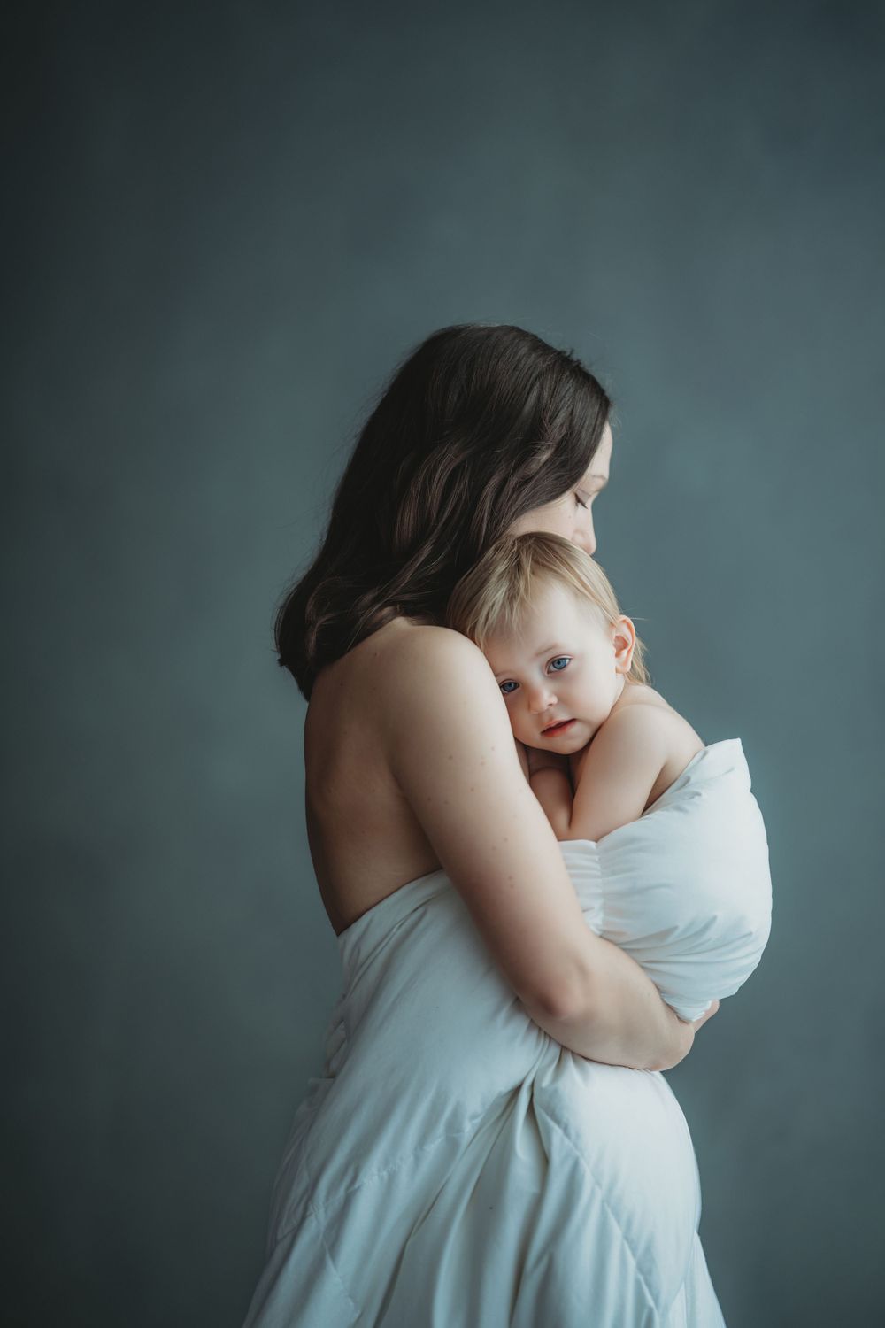 Intimate studio portrait of tender moment between parent and baby wrapped in soft white fabric against moody teal backdrop.
