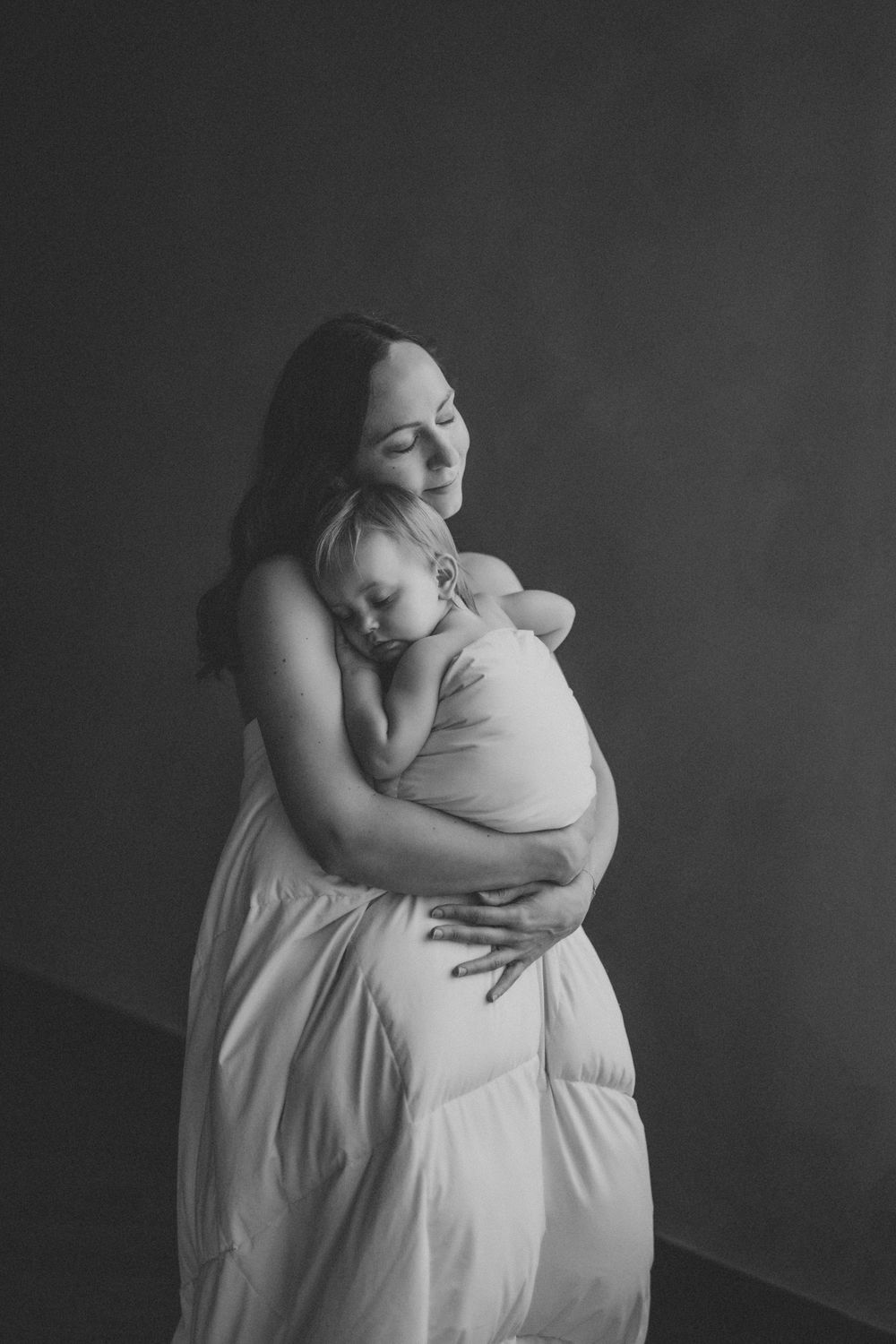 A tender black and white portrait of a mother and baby embracing in soft lighting against a dark background.