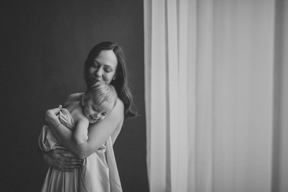 A graceful figure in a light dress smiles warmly while standing against a curtained wall in black and white.