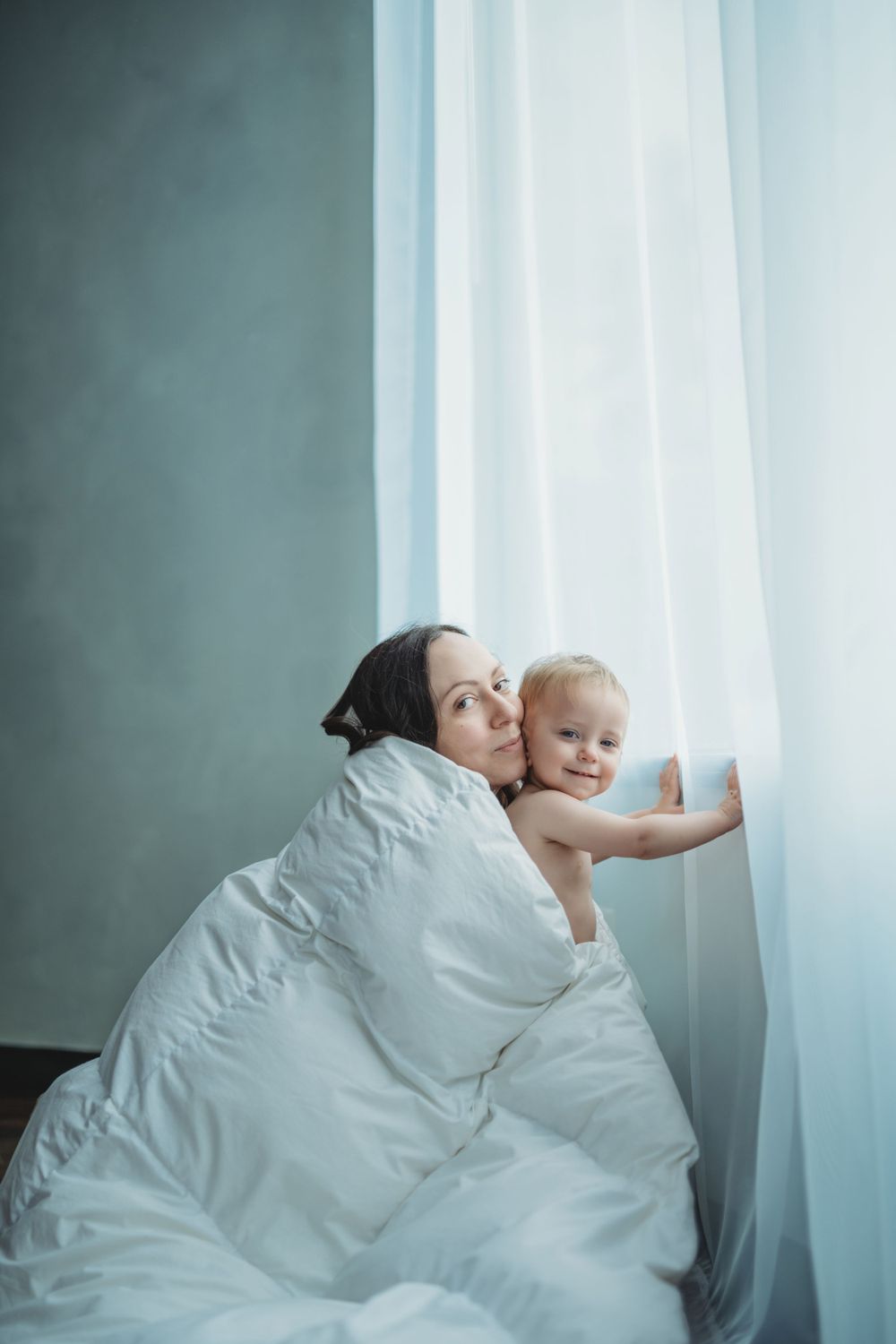 A parent and baby wrapped in white bedding look out through sheer curtains by a window.