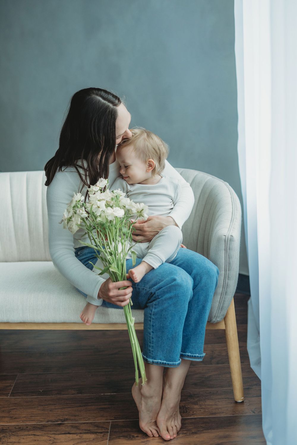 A tender moment on a white couch as parent and child share flowers in a blue-walled room with hardwood floors.