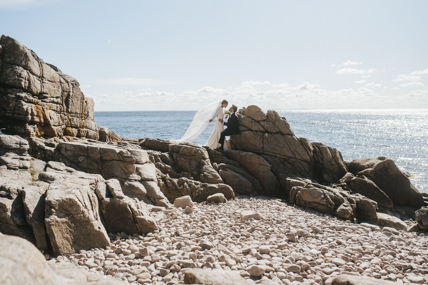 A figure in a flowing white dress stands gracefully on rugged coastal rocks overlooking a serene ocean view.