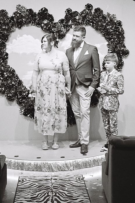 Black and white photo of a family posing together under a floral arch at a special occasion.