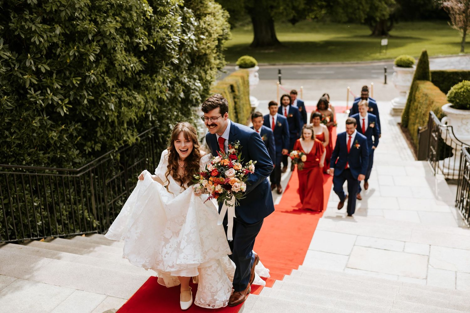 Wedding party walks down outdoor stairs in a formal procession during a sunny day at an elegant venue. Cabra Castle