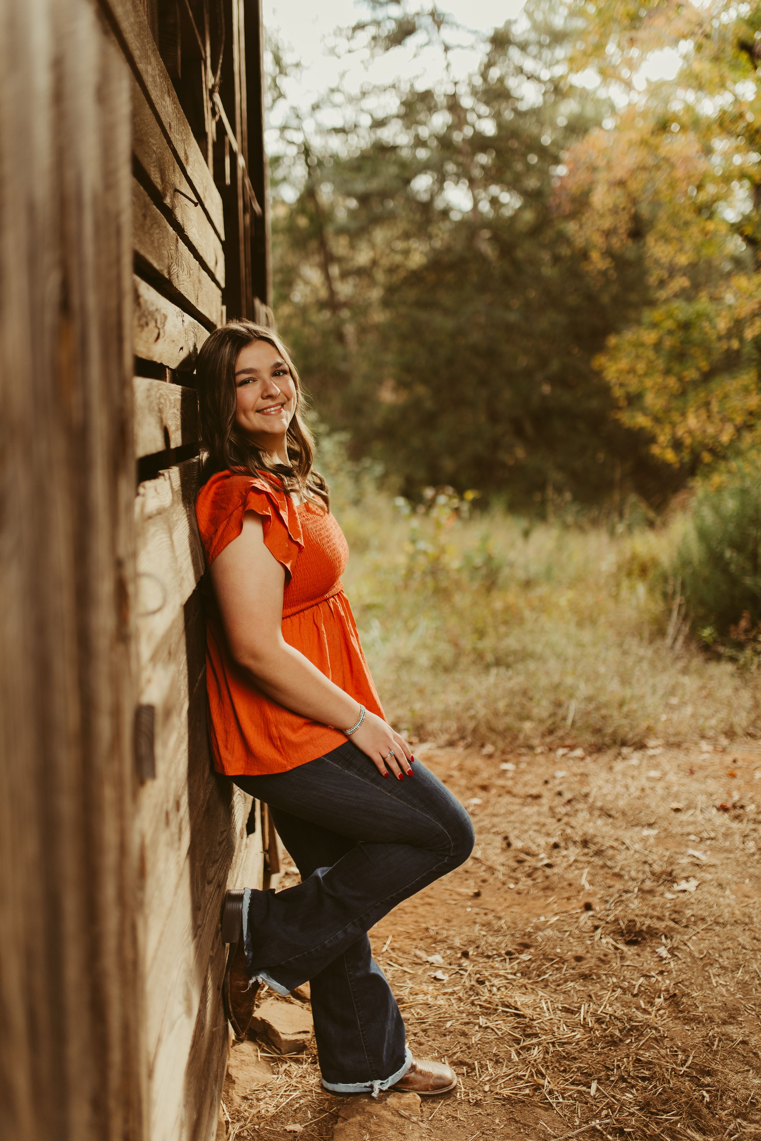 A smiling young woman in an orange top and jeans leans against a rustic wooden wall in golden sunlight.