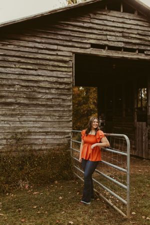 Young woman in orange top leaning on metal gate beside rustic wooden barn in autumn setting.