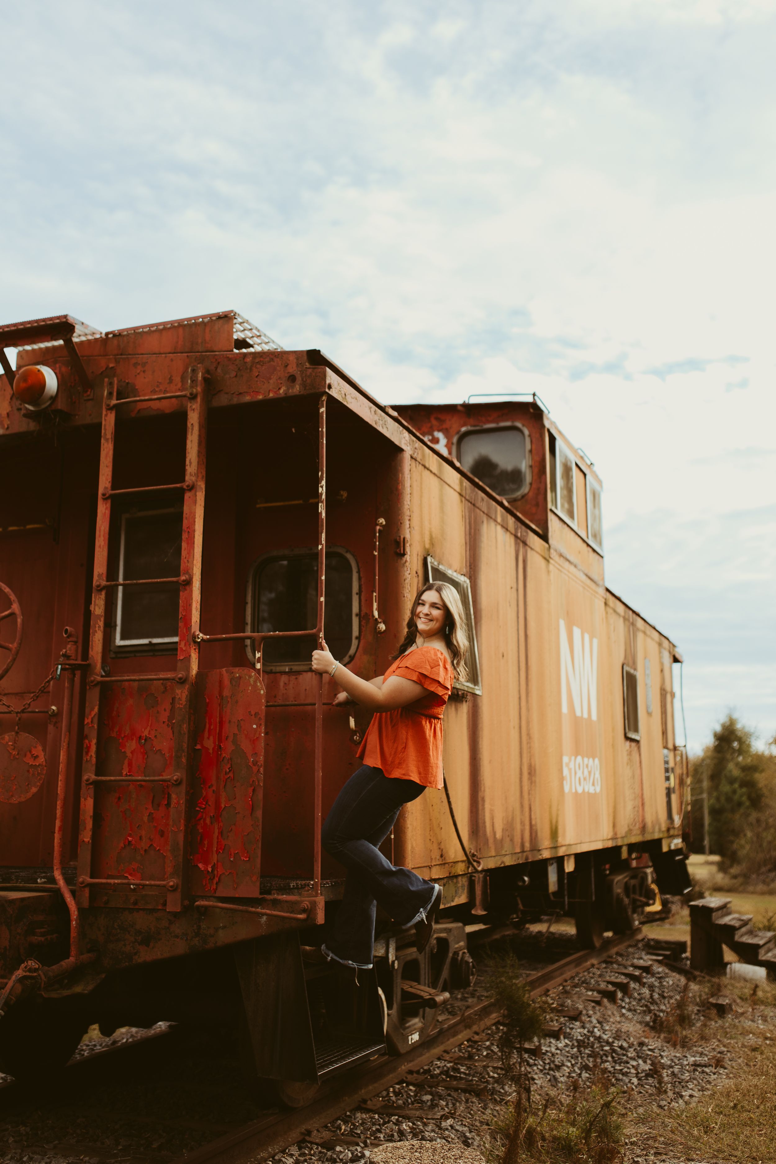 Young woman in orange top posing on steps of vintage red Norfolk Western caboose on railroad tracks.