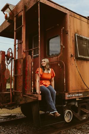 Smiling young woman in orange top sitting on vintage red train caboose steps on sunny day.
