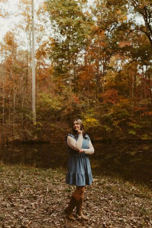 Young woman in blue denim dress and cowboy boots standing among vibrant autumn forest foliage.